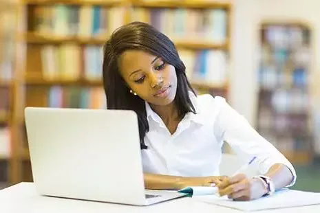 Woman in a library working on a laptop taking notes in a notebook